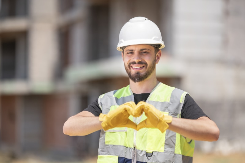 How to Join the HVAC Industry. Architect with white helmet and reflective vest is working at construction. He is smiling and looking at camera in front of a construction. He is making a heart shape.